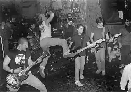 SSD playing live on a packed club stage, the singer leaping into the air mid-song beside the guitarists and bassist.